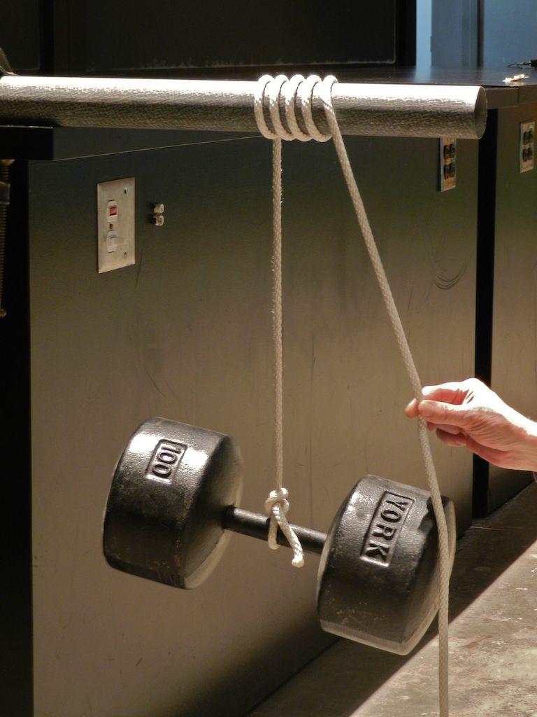 A rope tied to a 100 lb dumbell is wrapped around a post five times, and a person is holding the other end with finger and thumb.