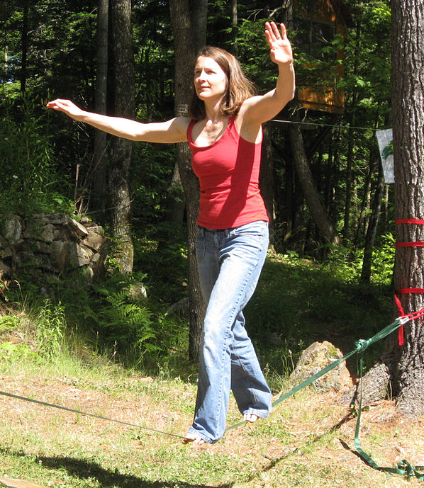 A woman walking across a slackline stretched between two trees. Her arms are outstretched for balance.